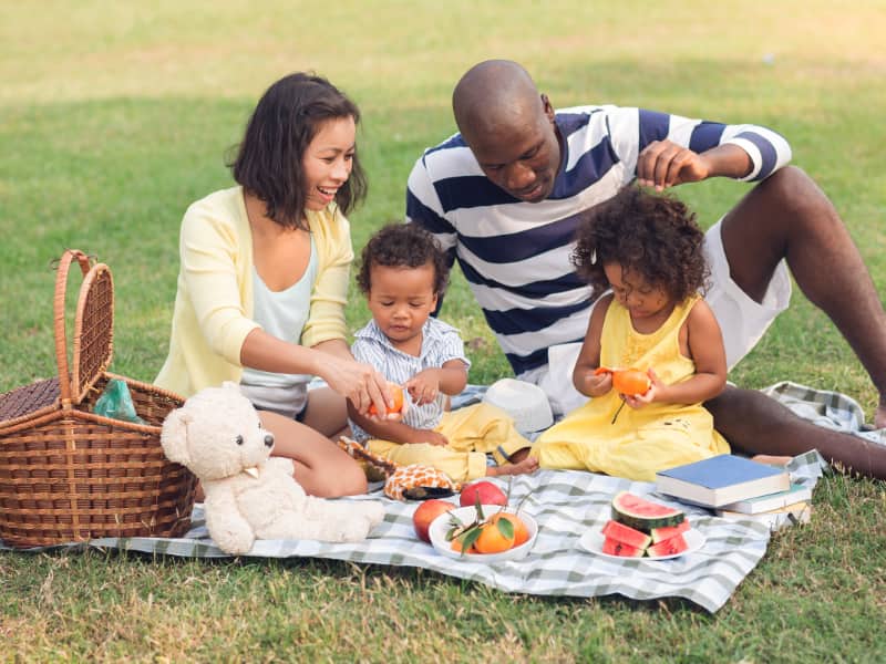 A multi-racial family with two young children sits down on a blanket having a picnic